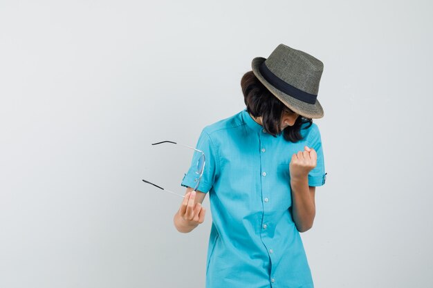 Foto gratuita giovane donna che mostra il gesto del vincitore mentre si tengono gli occhiali in camicia blu, cappello