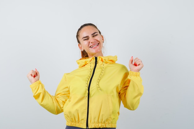 Молодая леди показывает жест победителя в желтой куртке и выглядит удачливым, вид спереди.