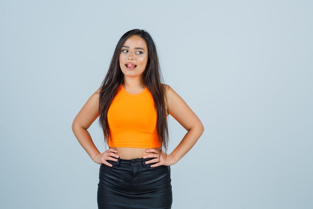 Free photo young lady in singlet, mini skirt holding hands on waist, sticking out tongue and looking cute , front view.