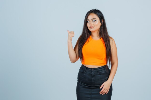 Young lady in singlet, mini skirt pointing back with thumb and looking confident , front view.