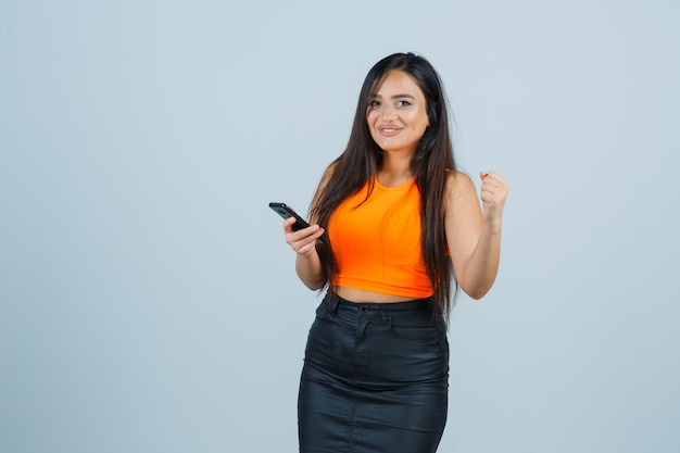 Young lady in singlet, mini skirt raising fist while looking at camera and looking blissful , front view.