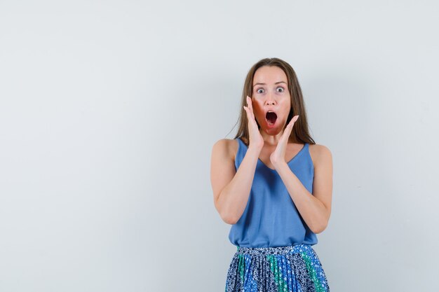 Young lady in singlet, skirt holding hands near open mouth and looking surprised , front view.