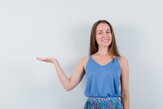 Young lady in singlet, skirt spreading palm aside and looking cheerful , front view.