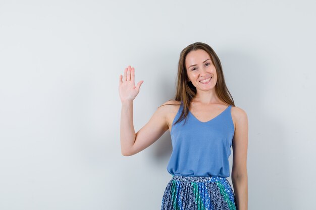 Young lady in singlet, skirt waving hand and looking cheerful , front view.