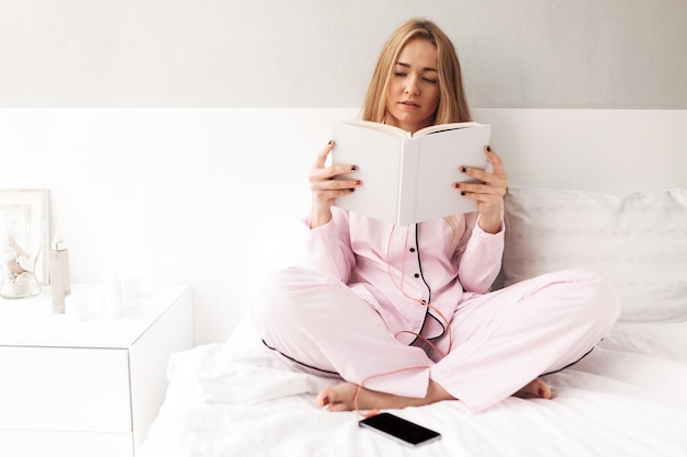 Барышня в пижаме сидит на кровати и читает книгу с мобильным телефоном рядом с домом