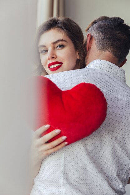 Young lady smiling and holding heart toy while hug her husband at home