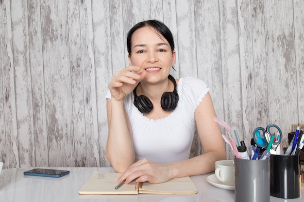 Free photo young lady smiling in white shirt drinking coffee listening to music in black earphones writing down notes on grey wall