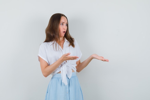 Young lady spreading her open palms aside for showing something in white blouse,blue skirt and looking dumbfounded