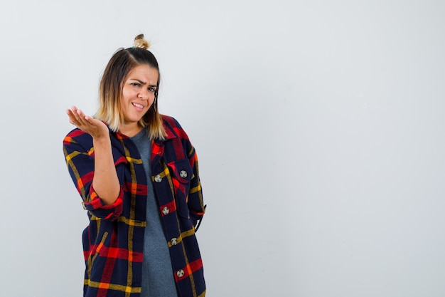 Young lady spreading palm in casual checked shirt and looking displeased , front view.