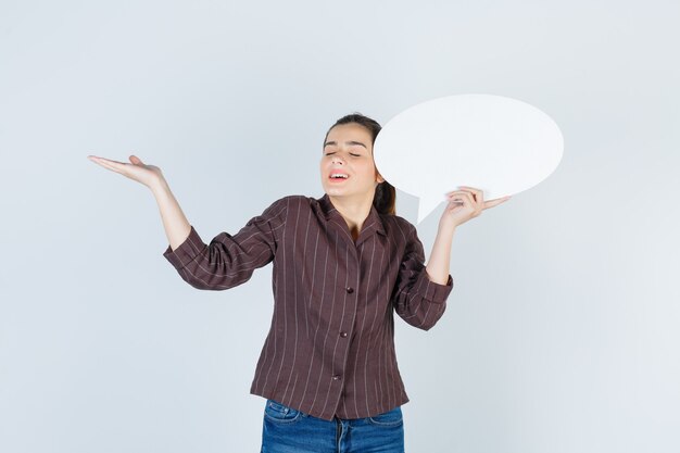 Young lady spreading palm, holding paper poster in shirt, jeans and looking happy , front view.