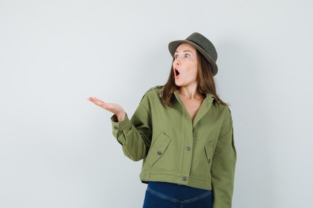 Young lady spreading palm in jacket pants hat and looking surprised