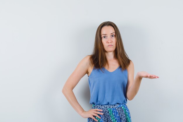 Young lady spreading palm in singlet, skirt and looking confused