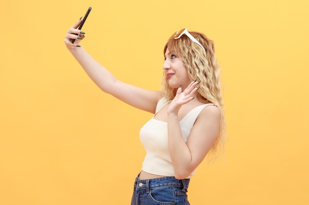 Девушка стоит на оранжевом фоне и делает селфи со своим телефоном