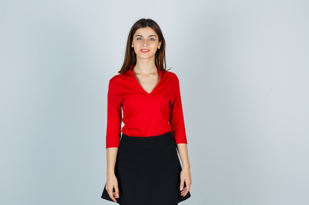 Девушка стоит прямо, позирует в красной блузке, черной юбке и выглядит весело