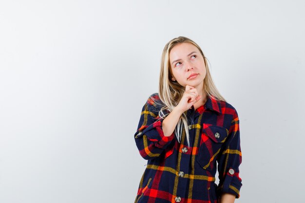 Young lady standing in thinking pose in checked shirt and looking hesitant , front view.