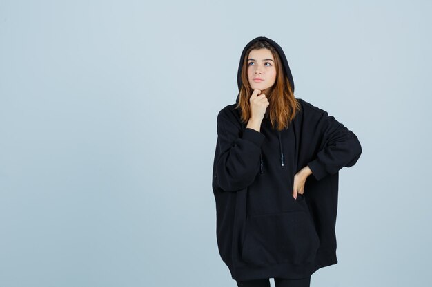 Young lady standing in thinking pose while keeping hand on waist in oversized hoodie, pants and looking thoughtful. front view.