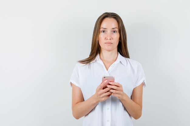 Young lady standing while holding phone in white blouse and looking serious