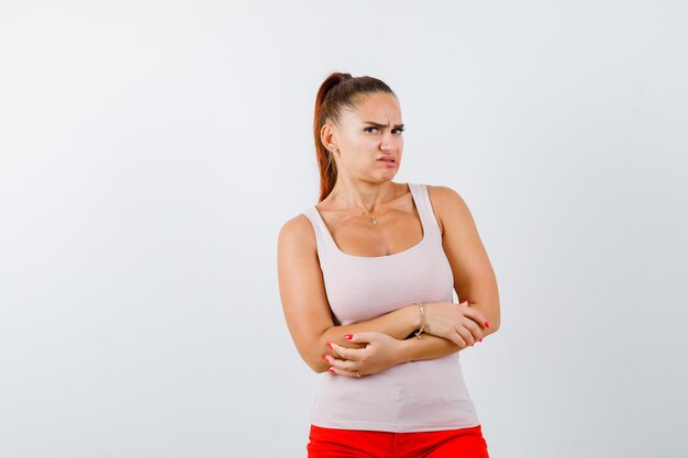 Young lady standing with crossed arms in beige tank top and looking dissatisfied. front view.