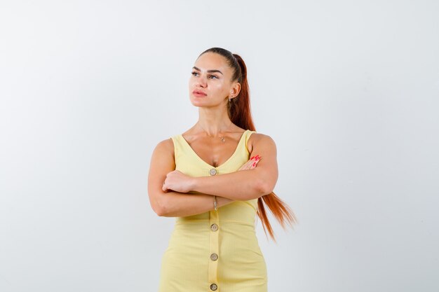 Young lady standing with crossed arms, looking away in yellow dress and looking pensive , front view.