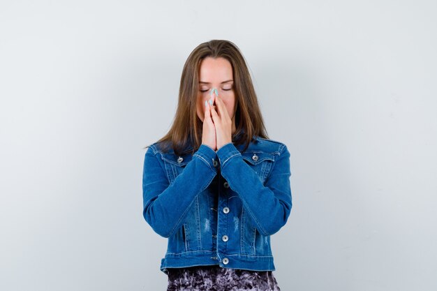 Young lady stands in praying gesture in blouse, denim jacket and looking hopeful , front view.