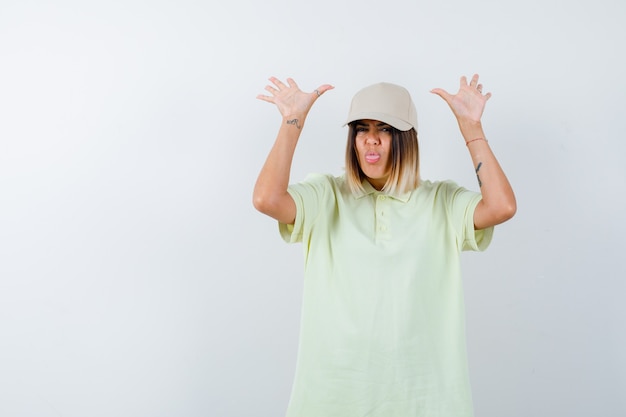 Young lady sticking out tongue while showing palms in surrender gesture in t-shirt, cap and looking helpless. front view.