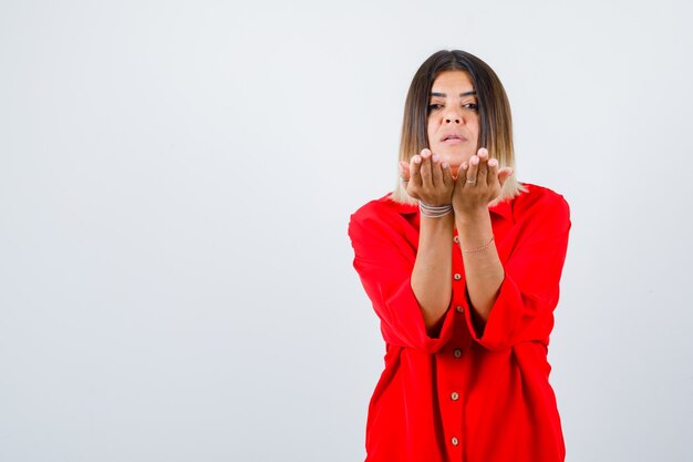 Young lady stretching cupped hands in red oversize shirt and looking confident , front view.
