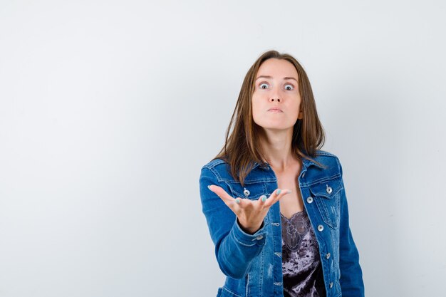 Young lady stretching hand in questioning gesture in blouse, denim jacket and looking angry , front view.