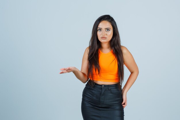 Young lady stretching hand in questioning gesture in singlet, mini skirt and looking serious , front view.