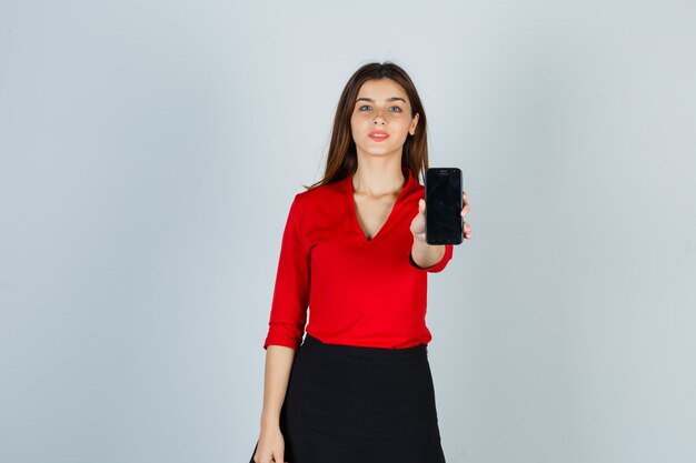 Young lady stretching hand to show mobile phone in red blouse, skirt and looking confident