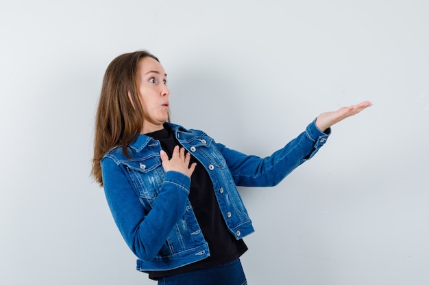 Young lady stretching hand to show something in blouse, jacket and looking puzzled. front view.