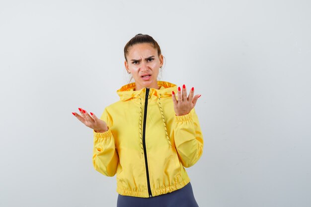 Young lady stretching hands in questioning gesture in yellow jacket and looking serious , front view.