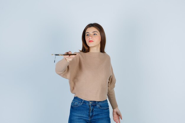 Young lady stretching mobile phone in beige sweater, jeans and looking serious. front view.