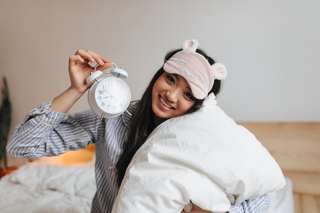 Young lady in striped pajamas keeps alarm clock Girl in sleep mask hugging pillow