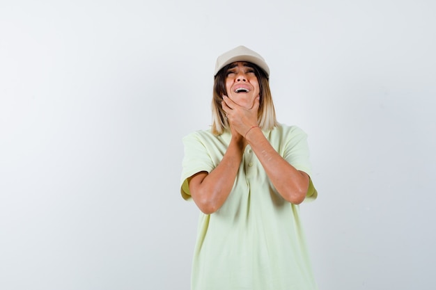 Young lady in t-shirt, cap holding hands under chin and looking offended , front view.