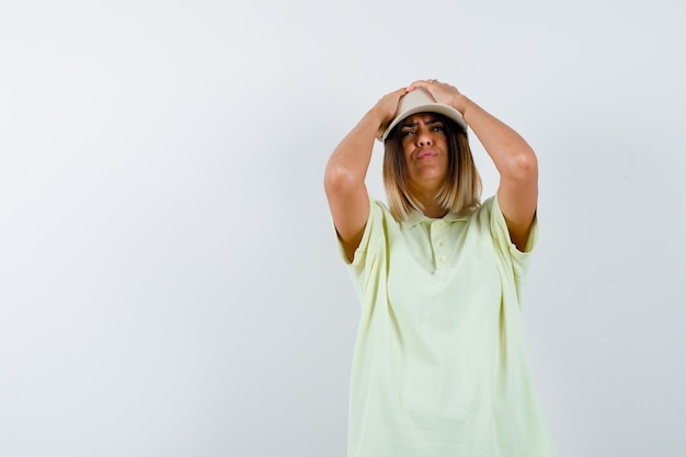 Young lady in t-shirt, cap holding hands on head and looking forgetful , front view.