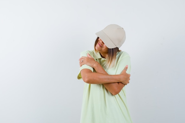 Free photo young lady in t-shirt, cap hugging herself and looking cute , front view.