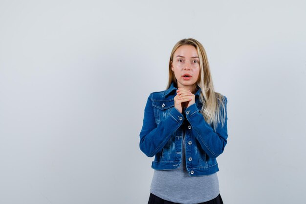 Young lady in t-shirt, denim jacket, skirt holding clasped hands near chest and looking attractive