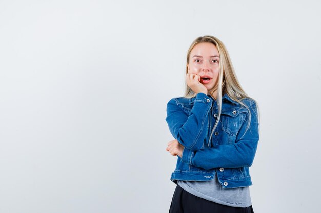 Young lady in t-shirt, denim jacket, skirt leaning chin on hand and looking amazed