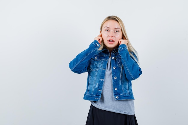 Young lady in t-shirt, denim jacket, skirt plugging ears with fingers and looking annoyed