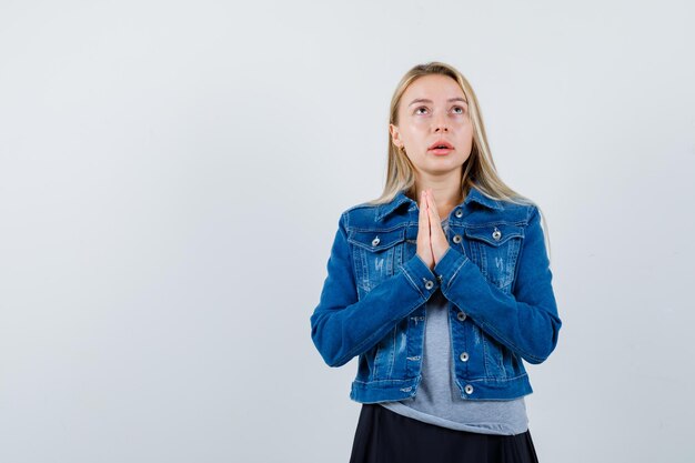 Young lady in t-shirt, denim jacket, skirt showing praying gesture and looking hopeful