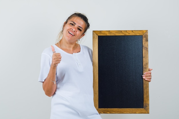 Young lady in t-shirt holding blackboard showing thumb up and looking cheerful  