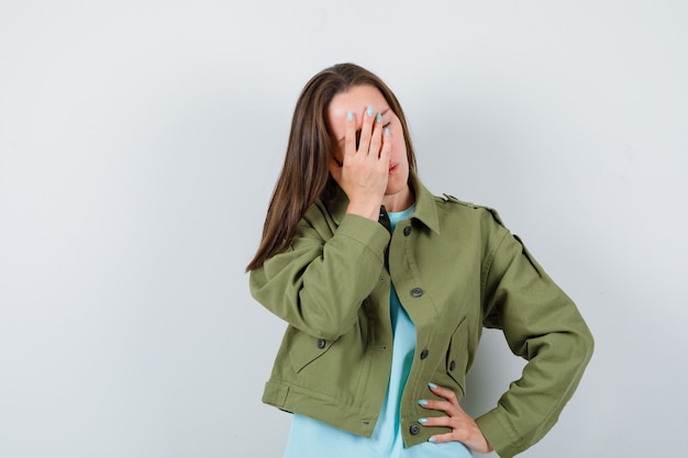 Free photo young lady in t-shirt, jacket covering face with hand while keeping hand on hip and looking forgetful , front view.