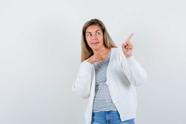 Young lady in t-shirt, jacket pointing at upper right corner and looking curious , front view.