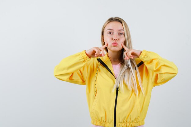 Young lady in t-shirt, jacket pressing blown cheeks with fingers and looking cute