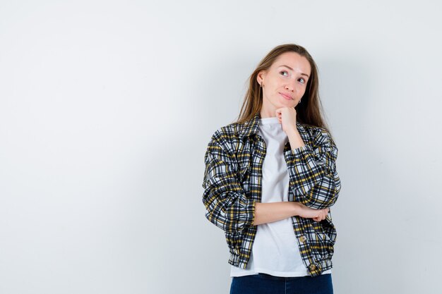 Young lady in t-shirt, jacket propping chin on hand and looking dreamy , front view.