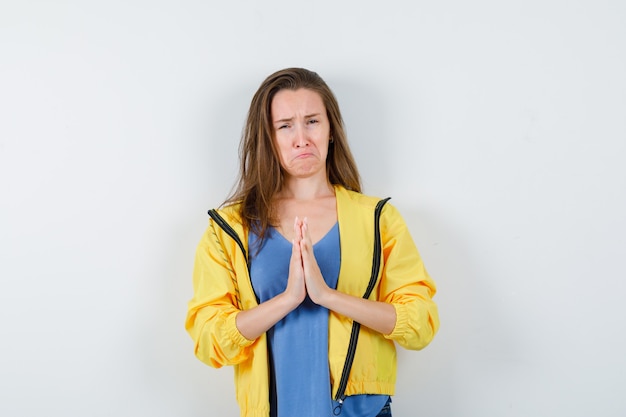 Giovane donna in t-shirt, giacca che mostra il gesto di namaste e sembra triste, vista frontale.