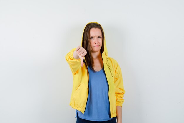 Young lady in t-shirt, jacket showing thumb down and looking displeased, front view.