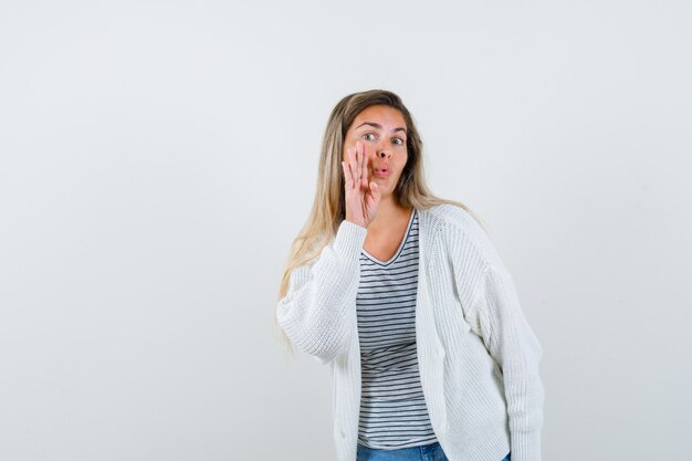 Young lady in t-shirt, jacket telling secret with hand near mouth and looking curious , front view.