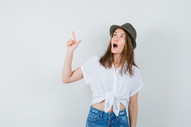 Young lady in t-shirt, jeans, hat pointing up and looking amazed , front view.
