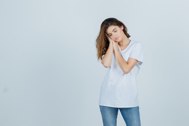 Young lady in t-shirt, jeans leaning on hands as pillow and looking sleepy , front view.